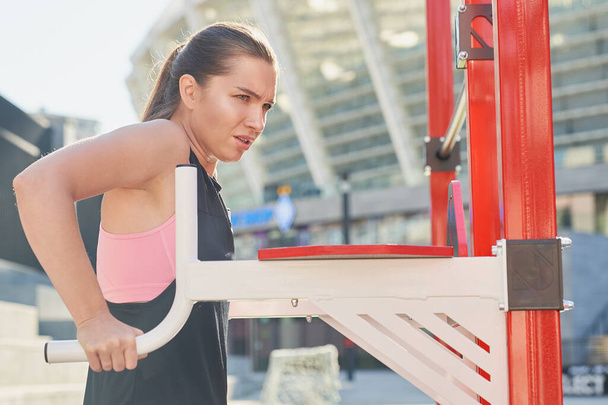 Serious, tense, young girl, athlete, in a T-shirt and top, does strength exercises with the help of a simulator on a street sports field, sports in the summer on the street. - Photo, Image