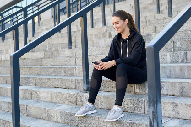 Stylish cute modern young woman in black sportswear, with long dark hair tied in a ponytail, sits on the steps outdoors and counts calories on her smartphone. Sports, fitness and technology. - Foto, Imagen