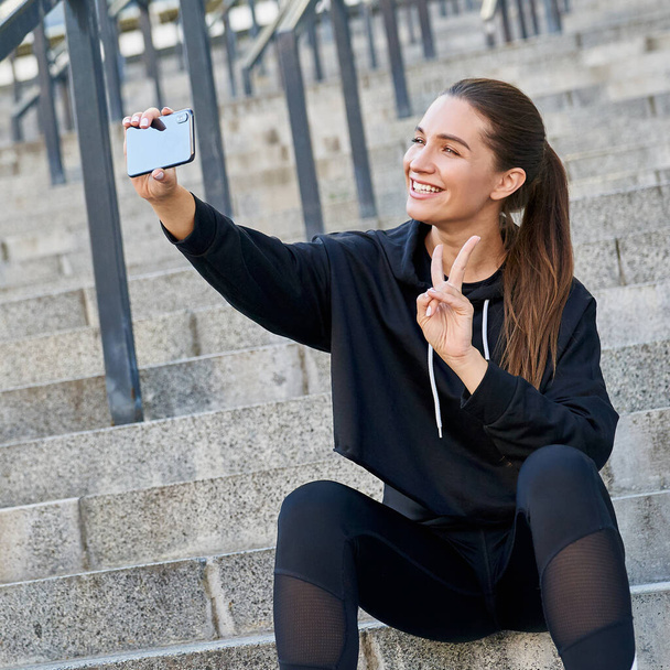 Slim beautiful modern young girl athlete, fitness lady, in black sportswear, sits on the gray steps outdoors and takes a selfie and shows the peace sign. Sports, fitness and technology. - Fotoğraf, Görsel
