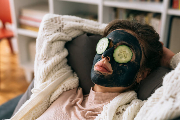 woman with cosmetic face mask and cucumber on her eyes, relaxing on sofa at her home - Fotografie, Obrázek