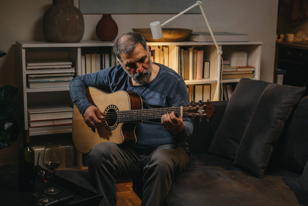 man van middelbare leeftijd speelt akoestische gitaar thuis, 's avonds scene - Foto, afbeelding