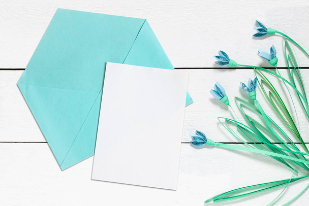 Tarjeta en blanco o maqueta de invitación, sobre azul y gotas de nieve en técnica de extracción sobre fondo de madera blanca. Concepto primavera - Foto, Imagen