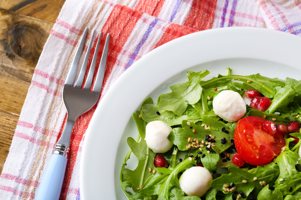 Green salad made with  arugula, tomatoes, cheese mozzarella balls and sesame  on plate, on wooden background - Photo, image
