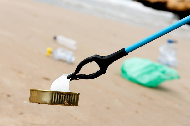 qualcuno raccoglie un cibo usato può, con un estensore di portata, dalla sabbia vicino alla riva di una spiaggia solitaria, dove ci sono altri rifiuti, come una bottiglia di plastica o un sacchetto di plastica verde - Foto, immagini