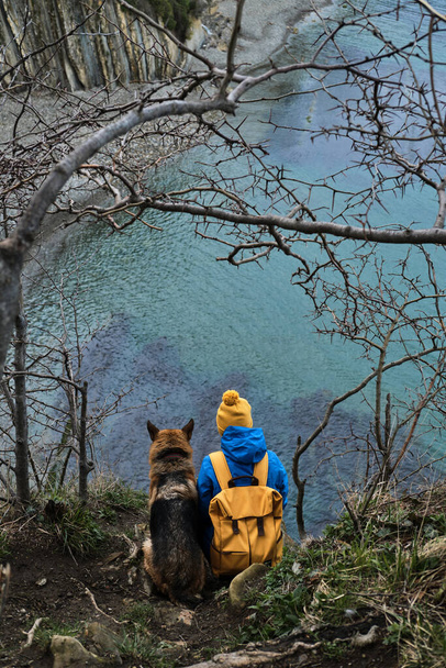 Chien avec propriétaire sur la mer. Fille avec sac à dos jaune et chapeau se trouve sur le bord de la falaise surplombant l'océan bleu à côté de l'ami berger allemand noir et rouge. Promenade et voyage avec chien. - Photo, image
