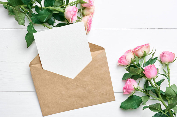 Greeting card mockup with envelope and fresh pink roses on white wooden background, mockup with copy space - Foto, Imagen