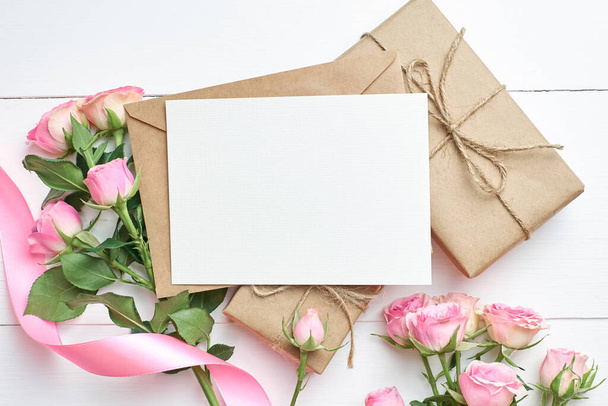Greeting card mockup with fresh roses and gift boxes on white wooden background, top view, copy space - Photo, image