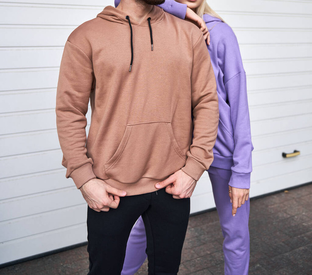 Blonde girl is standing in blue sport outfit. Man wears brown hoodie and black pants. Couple is wearing street matching outfit - Photo, Image