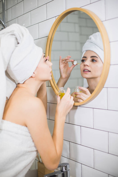Jeune femme en serviette blanche appliquer de l'huile hydratante sur la peau du visage. - Photo, image