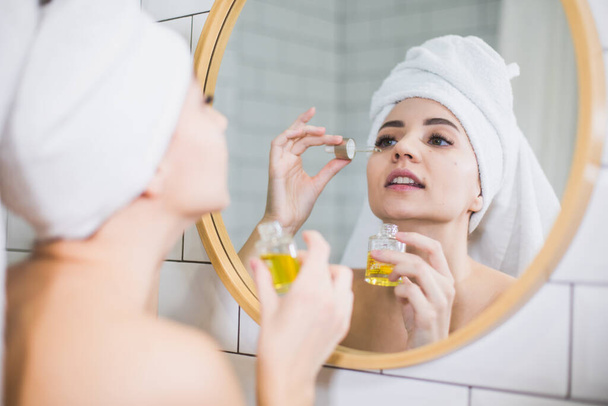 Jeune femme en serviette blanche appliquer de l'huile hydratante sur la peau du visage. - Photo, image