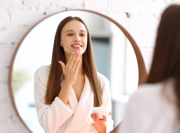 Joyeux jeune femme en peignoir appliquer de la crème cosmétique sur le visage et profiter de la douceur de la peau lors d'une séance de beauté spa à la maison - Photo, image