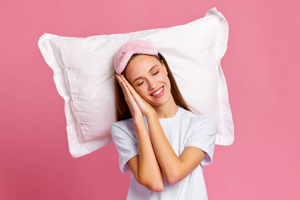 Delighted young female clasping hands under cheek and smiling while sleeping on soft pillow against pink background - Фото, изображение