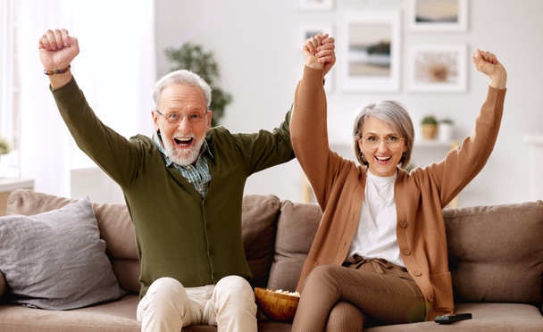 Iloinen vanhempi pari aviomies ja vaimo juhlivat maalia istuessaan sohvalla olohuoneessa ja katsoessaan jalkapallo-ottelua televisiosta, onnellinen kypsä perhe pitää hauskaa yhdessä viettäessään viikonlopun kotona. - Valokuva, kuva