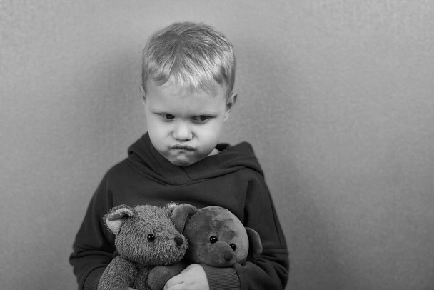 Ein dreijähriger kaukasischer Junge sitzt allein auf dem Boden und umarmt Teddybären, das Thema Kindesmissbrauch, Schwarz-Weiß-Foto. - Foto, Bild