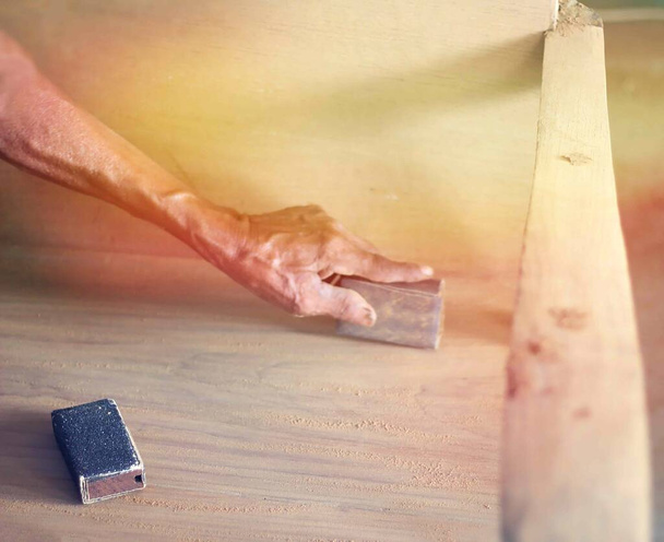 Η διαδικασία άλεσης του ξύλου με γυαλόχαρτο. Ο γερο-ξυλουργός δουλεύει με ξύλο. Λαβή γυαλόχαρτου. Ηλιακό φως. Ξύλινο μπράτσο. - Φωτογραφία, εικόνα