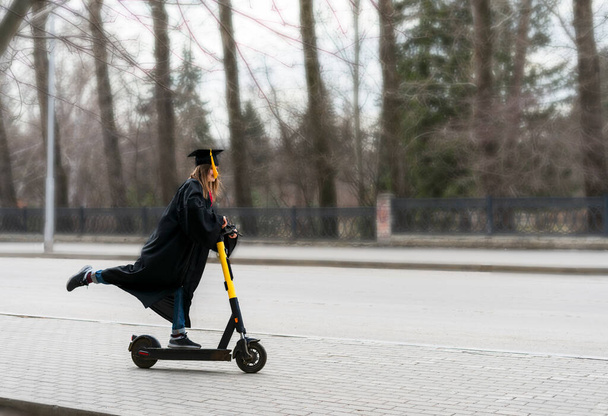 Felice Giovane donna in abito da laurea nero e tappo nero graduazione con nappa gialla cavalcando uno scooter elettrico in fondo alla strada dopo la laurea all'università - Foto, immagini