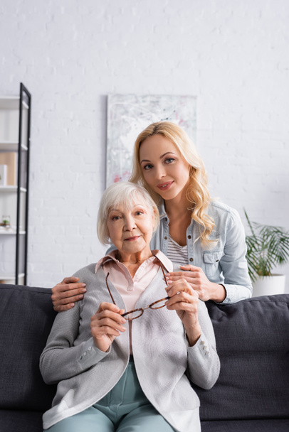 Woman hugging elderly mother with eyeglasses on couch  - Valokuva, kuva