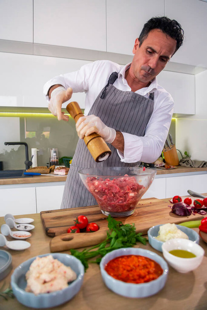 De chef-kok voegt de kruiden toe in een transparante glazen kom met vlees, ui en spek, terwijl aan de zijkant van het werkoppervlak groenten, plakjes spek, specerijen en keukengerei staan.. - Foto, afbeelding
