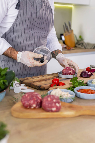 Un primo piano di uno chef che modella carne per hamburger in una pressa su una tavola di legno in cucina. Ci sono varie spezie, verdure, così come piccole ciotole da cucina intorno al piano di lavoro. - Foto, immagini