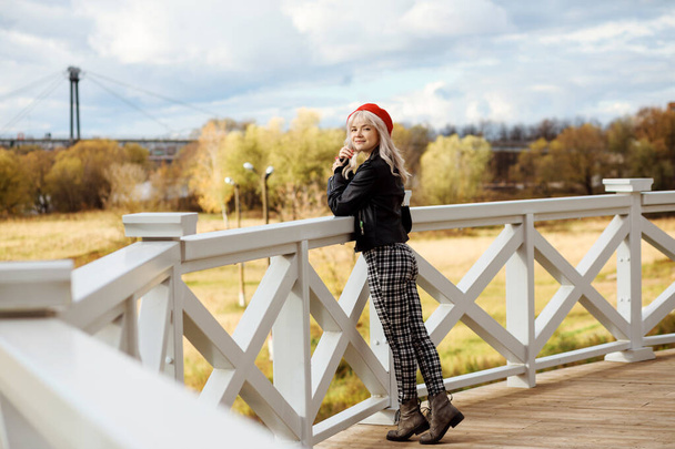 Full length portrait of attractive blonde woman outdoors in autumn. Fashionable woman is wearing checkered trousers, black leather jacket, red beret. Casual female street style.  - Zdjęcie, obraz