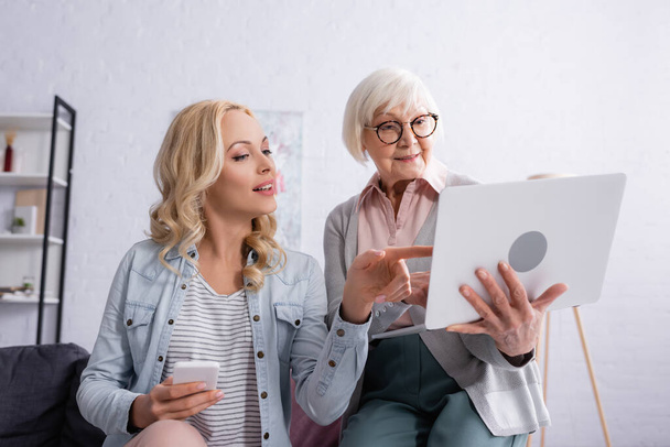 Smiling woman with smartphone pointing at laptop near senior mother at home  - Photo, image