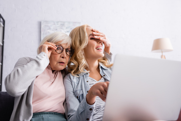 Excited woman covering eyes of daughter near blurred laptop at home  - Photo, Image