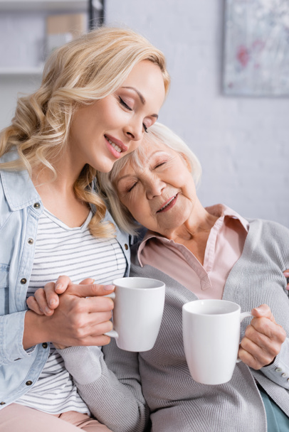 Smiling elderly woman holding cup while sitting near adult daughter  - Foto, immagini