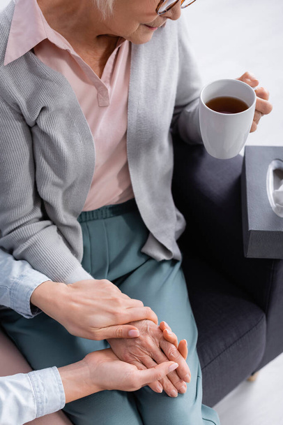 Vista recortada de la mujer mayor sosteniendo la taza de té cerca de la hija sosteniendo la mano  - Foto, Imagen