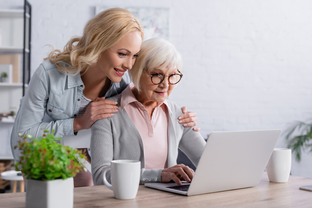 Smiling woman hugging mother using laptop near cups  - Photo, Image