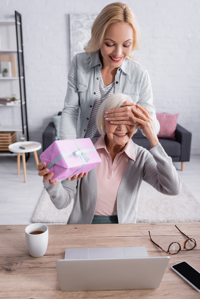 Улыбающаяся женщина закрывает глаза на мать, держа в руках ноутбук и чай на столе  - Фото, изображение