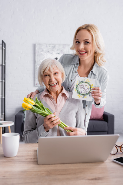 Lächelnde Seniorin hält Blumen neben Tochter mit Grußkarte neben Laptop  - Foto, Bild