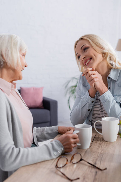 Positive woman talking to mother with cup of tea near eyeglasses  - Photo, Image