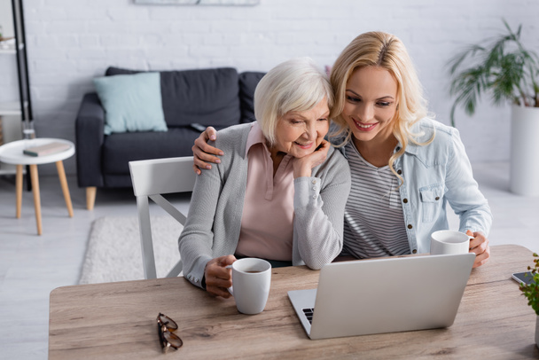Smiling woman hugging mother with cup near laptop on table  - Foto, Bild