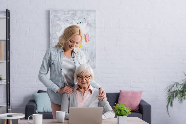 Sorrindo mulher de pé perto da mãe sênior usando laptop na sala de estar  - Foto, Imagem