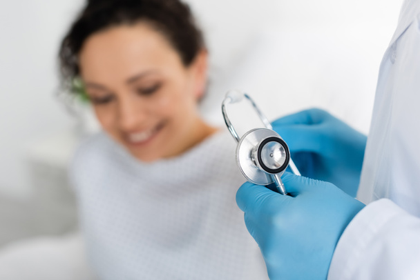 doctor en guantes de látex sosteniendo estetoscopio cerca de sonriente mujer afroamericana sobre fondo borroso - Foto, Imagen