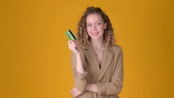 Cheerful young girl with curly hair Point hand on credit bank card showing thumb up like gesture isolated on yellow studio background. People lifestyle concept. - Video, Çekim