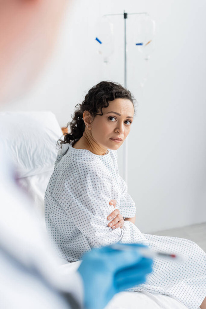 mujer afroamericana molesta sentada en la cama del hospital cerca del médico con termómetro en primer plano borroso - Foto, Imagen