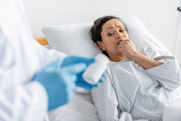 worried african american woman lying in hospital bed near blurred doctor in latex gloves pointing at container with medication - Φωτογραφία, εικόνα