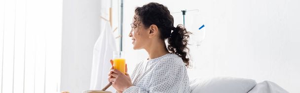 χαμογελαστή Αφροαμερικάνα που κοιτάζει αλλού ενώ κρατάει χυμό πορτοκάλι στο νοσοκομείο, πανό - Φωτογραφία, εικόνα