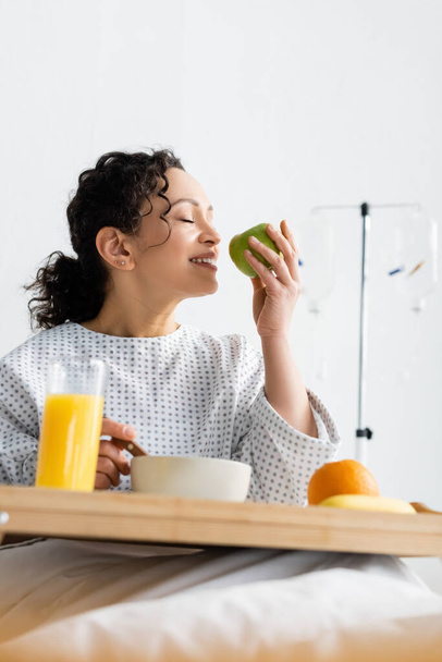 agradecida mujer afroamericana disfrutando del sabor de la manzana madura durante el desayuno en el hospital - Foto, Imagen