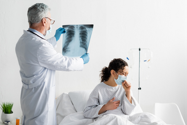 radiolog v lékařské masce drží plíce x-ray v blízkosti africké americké masky kašel na nemocniční posteli - Fotografie, Obrázek