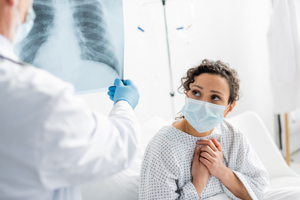 обеспокоенная африканская американка в медицинской маске рядом с рентгенологом в латексной перчатке держит рентген легких на размытом переднем плане - Фото, изображение