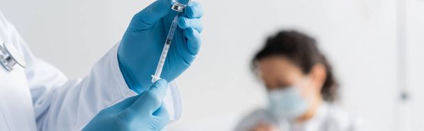 bijgesneden zicht op arts in latex handschoenen met spuit met vaccin in de buurt van Afro-Amerikaanse vrouw op wazige achtergrond, spandoek - Foto, afbeelding