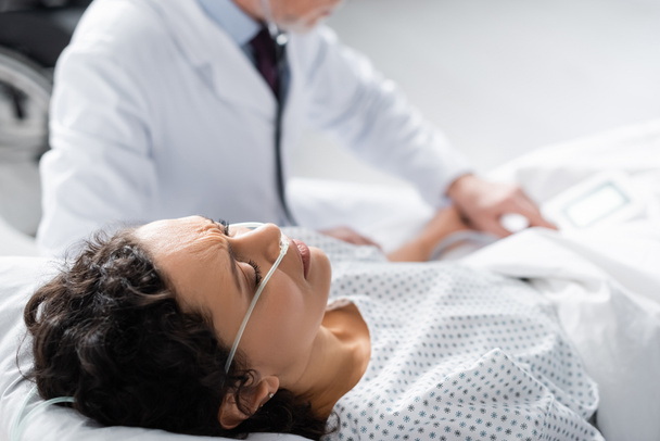 mujer afroamericana enferma con cánula nasal acostada en la cama cerca del médico sobre fondo borroso - Foto, imagen