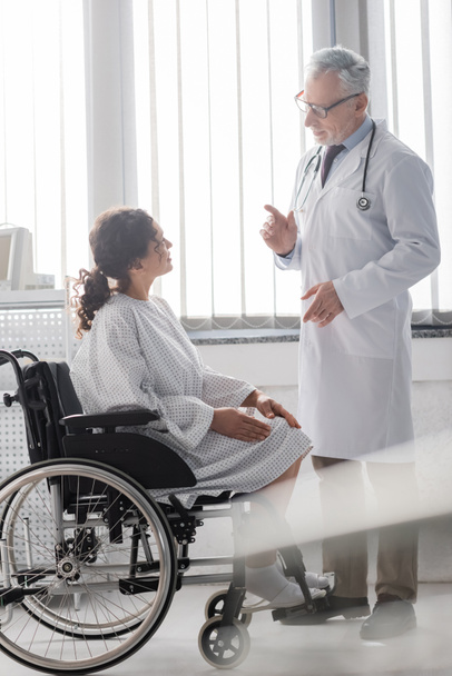 Dojrzały lekarz rozmawia z afrykańską Amerykanką siedzącą na wózku inwalidzkim na niewyraźnym pierwszym planie - Zdjęcie, obraz
