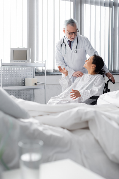 happy african american woman in wheelchair gesturing while talking to doctor near bed on blurred foreground - Photo, Image