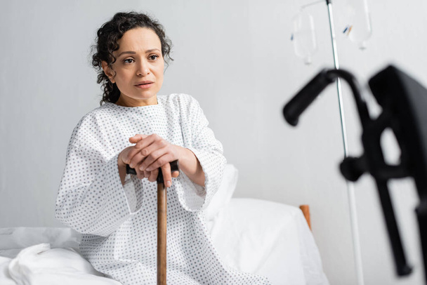 upset african american woman sitting on hospital bed with walking stick on blurred foreground - Fotó, kép