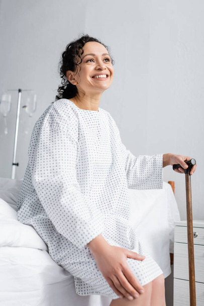 joyful african american woman touching knee while sitting on bed with walking stick - Photo, Image
