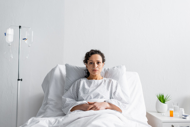 mujer afroamericana enferma mirando la cámara mientras está sentada en la cama del hospital cerca del mostrador de entrega - Foto, Imagen
