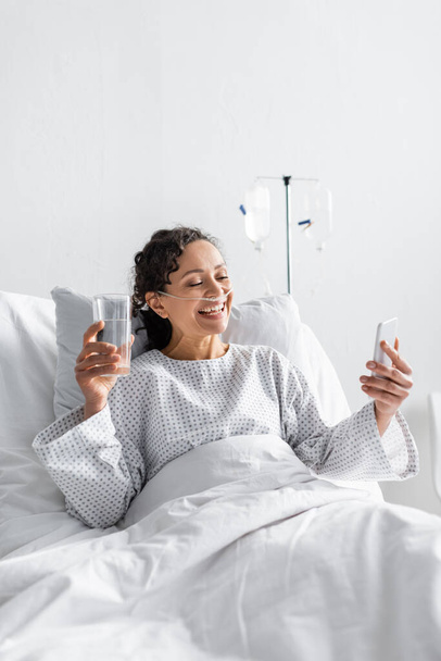 alegre mujer afroamericana mirando el teléfono inteligente mientras sostiene un vaso de agua en el hospital - Foto, imagen